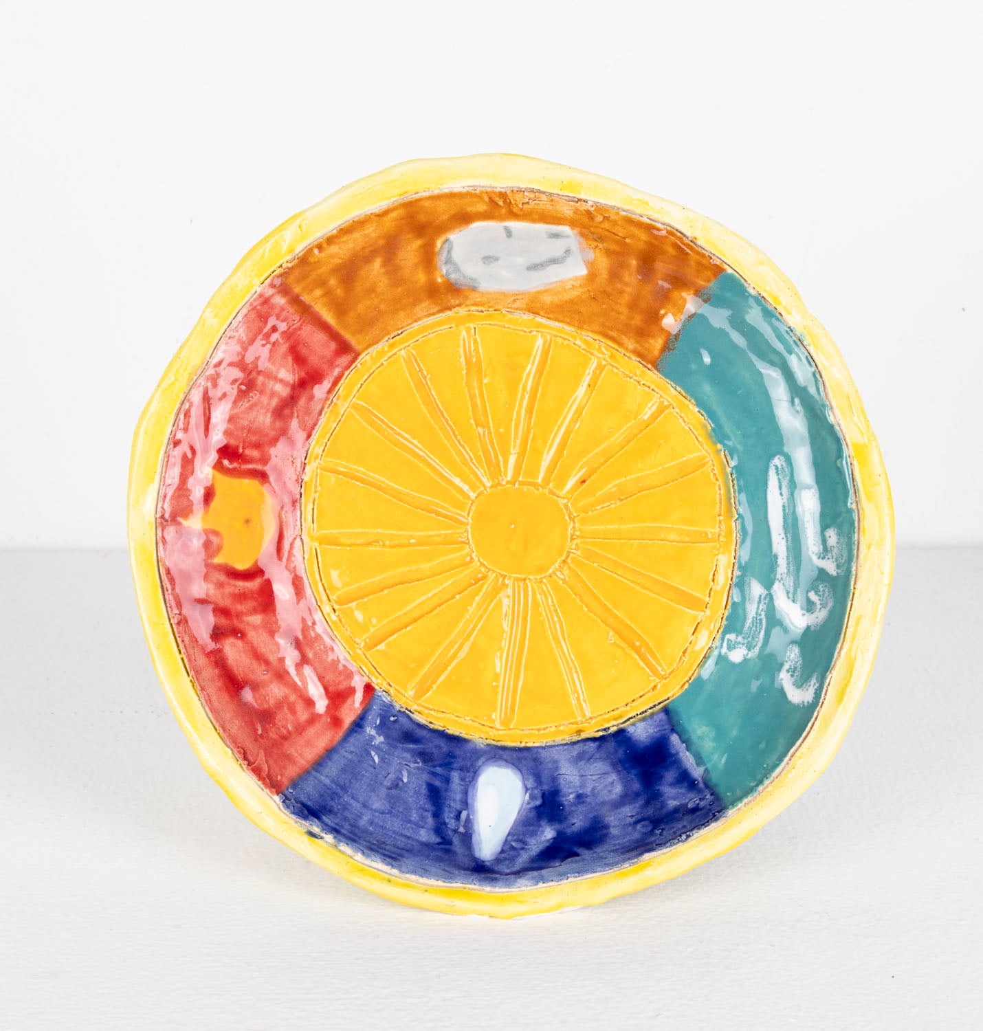 This is a ceramic sculpture of a plate, the plate has a sun carved into the center, with a yellow glaze. The perimeter of the plate is divided into sections, each section being a different color; red, brown, green and blue. The border of the plate is also a yellow glaze.
