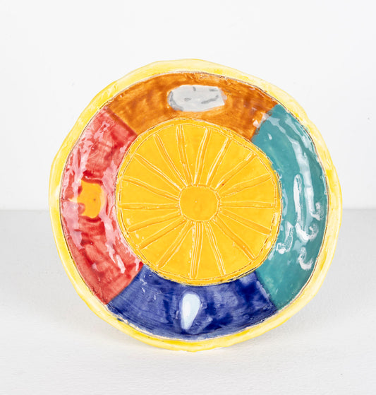 This is a ceramic sculpture of a plate, the plate has a sun carved into the center, with a yellow glaze. The perimeter of the plate is divided into sections, each section being a different color; red, brown, green and blue. The border of the plate is also a yellow glaze.
