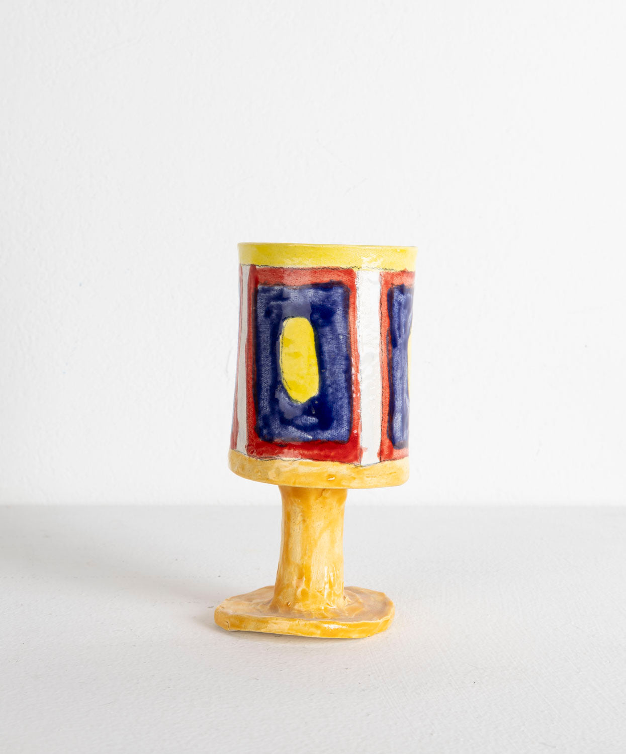 This is a ceramic sculpture of a chalice. The chalice is predominantly glazed in yellow, with a rectangular pattern created with blue, yellow and red glaze.