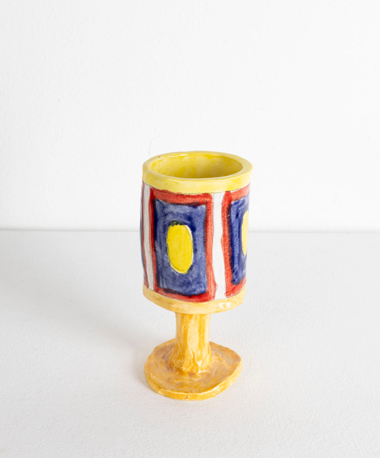 This is a ceramic sculpture of a chalice. The chalice is predominantly glazed in yellow, with a rectangular pattern created with blue, yellow and red glaze.