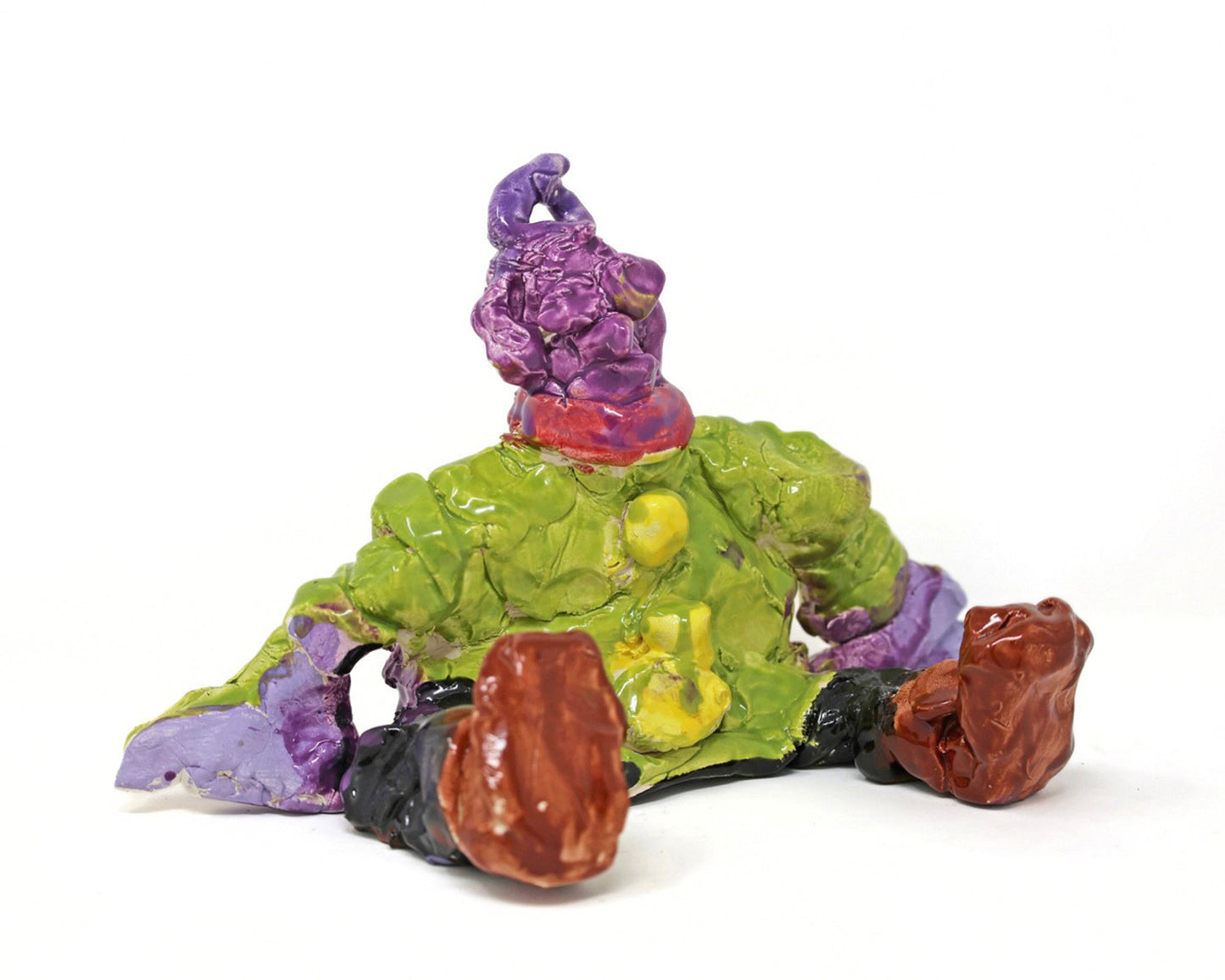 This ceramic sculpture depicts a figure that is sitting down. The figure has purple skin, and is wearing a green shirt with yellow buttons, a red scarf, black pants and brown shoes. 