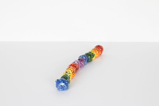 Colorful ceramic caterpillar sculpture made of connected rounded segments in rainbow colors with black spots. The caterpillar has a blue smiling face at the front and is displayed on a plain light gray surface against a neutral background.
