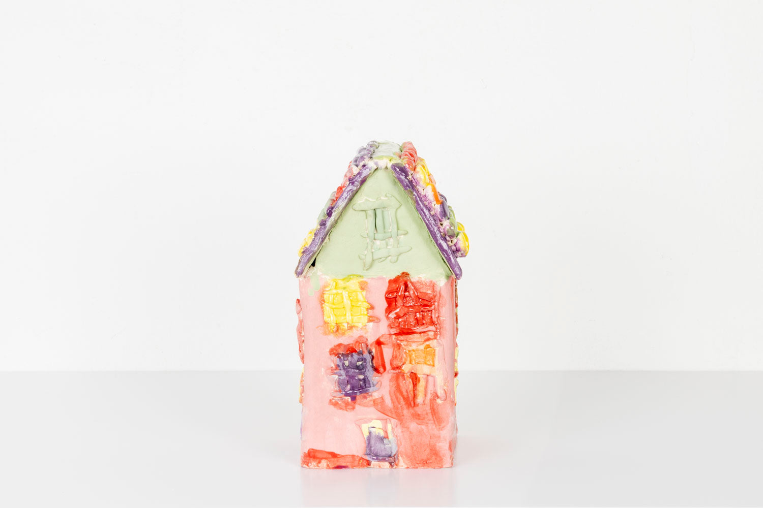 A small handmade ceramic house sculpture with a bright red body and a pale green roof, displayed against a plain white background. The house is decorated with colorful textured details, including pastel windows and flower-like accents in yellow, purple, and orange along the roofline. 