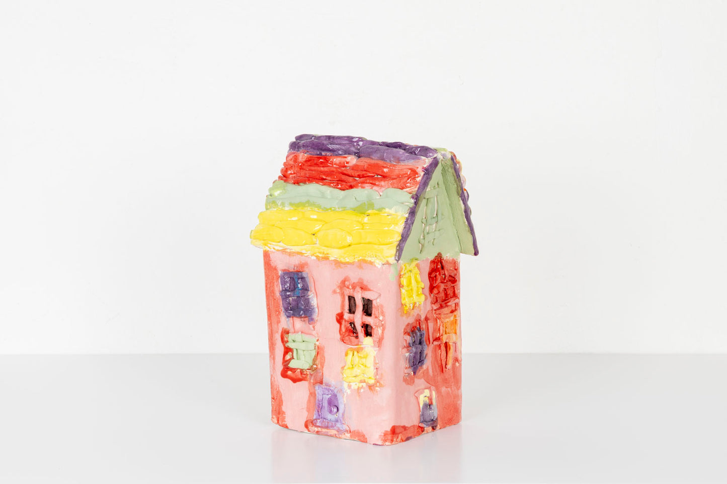 A small handmade ceramic house sculpture with a bright red body and a pale green roof, displayed against a plain white background. The house is decorated with colorful textured details, including pastel windows and flower-like accents in yellow, purple, and orange along the roofline. 