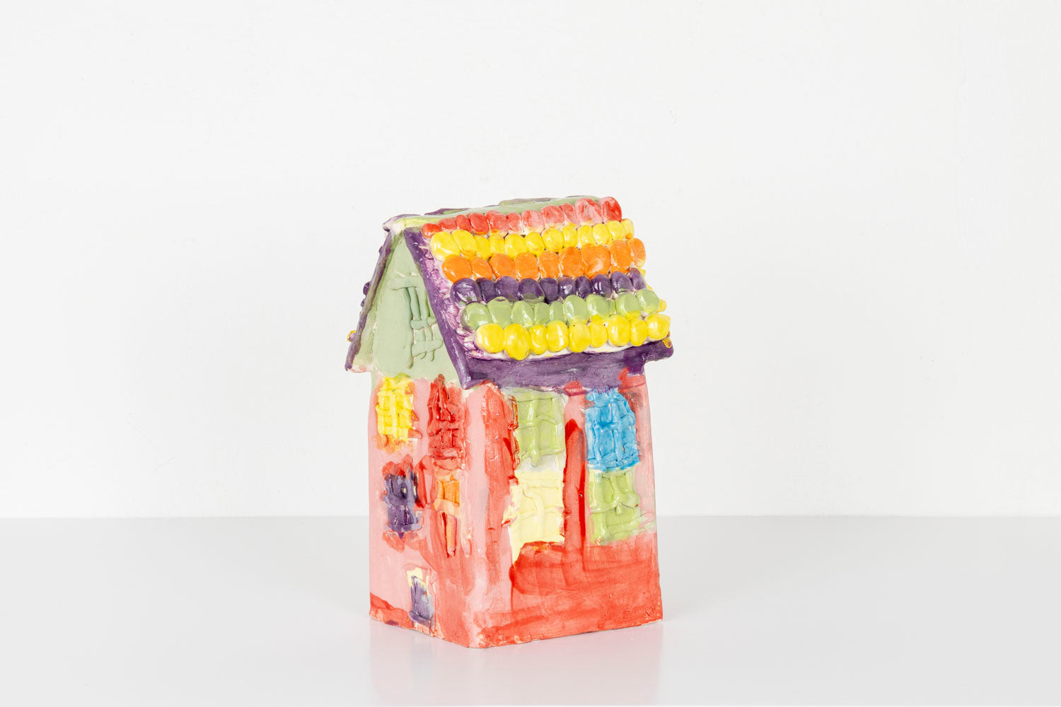 A small handmade ceramic house sculpture with a bright red body and a pale green roof, displayed against a plain white background. The house is decorated with colorful textured details, including pastel windows and flower-like accents in yellow, purple, and orange along the roofline. 