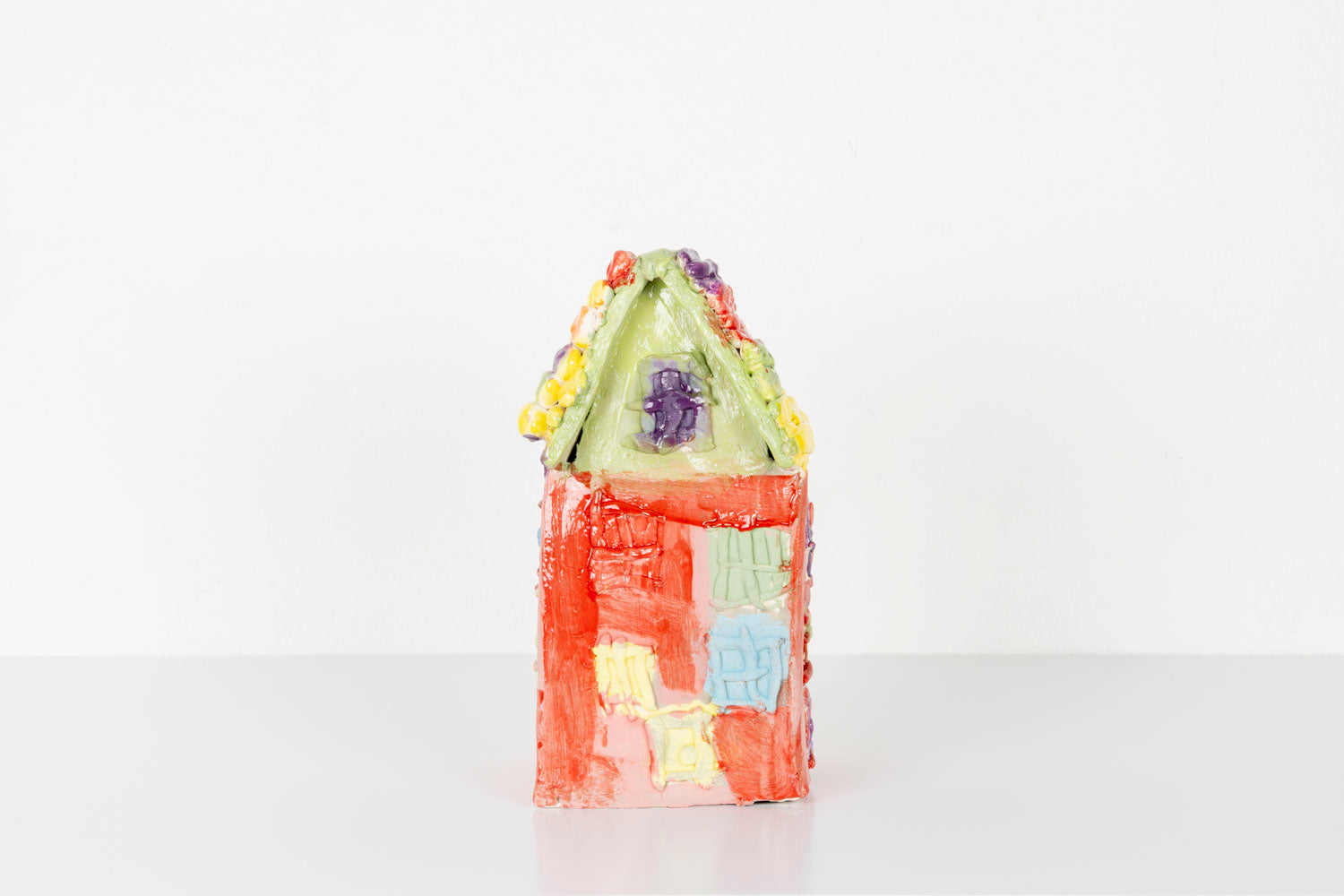 A small handmade ceramic house sculpture with a bright red body and a pale green roof, displayed against a plain white background. The house is decorated with colorful textured details, including pastel windows and flower-like accents in yellow, purple, and orange along the roofline. 