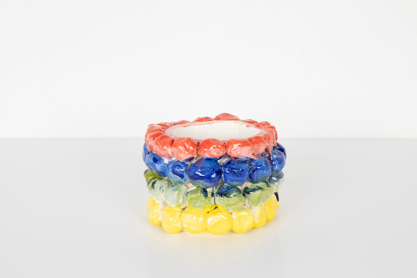 A small cylindrical ceramic vessel with a glossy surface, decorated with rows of rounded bubble-like shapes in bright colors. The top row is coral pink, followed by blue, pale green, and yellow bands. The piece sits on a white surface against a plain light background.