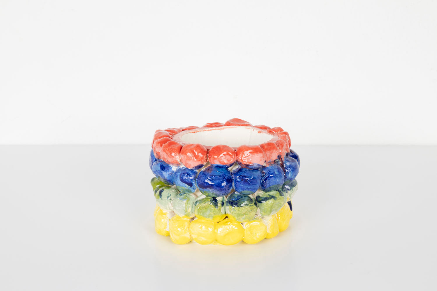 A small cylindrical ceramic vessel with a glossy surface, decorated with rows of rounded bubble-like shapes in bright colors. The top row is coral pink, followed by blue, pale green, and yellow bands. The piece sits on a white surface against a plain light background.