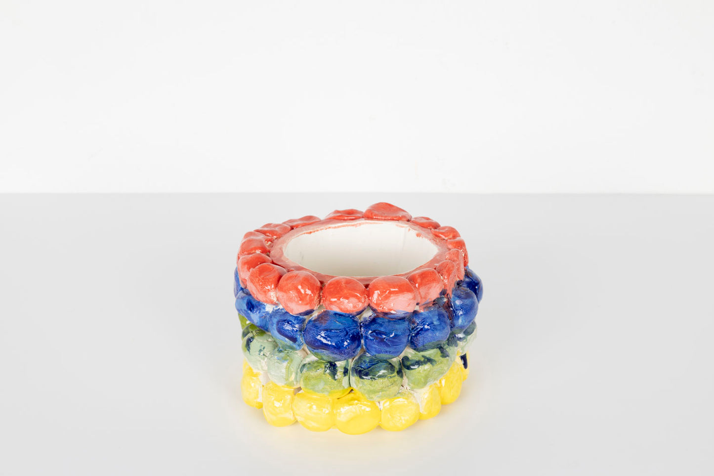 A small cylindrical ceramic vessel with a glossy surface, decorated with rows of rounded bubble-like shapes in bright colors. The top row is coral pink, followed by blue, pale green, and yellow bands. The piece sits on a white surface against a plain light background.