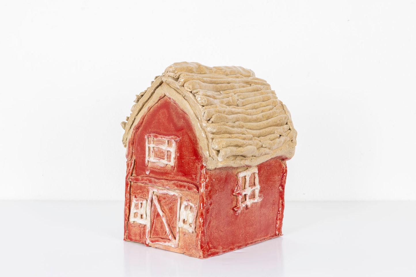 This is a ceramic sculpture of a house, glazed with vibrant hues of red and beige. The details of the house such as the windows and the doors are carved into the clay. This side view highlights the textures on the roof that contrasts the smooth surface of the rest of the house.