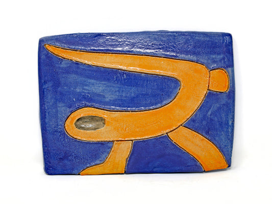 This ceramic piece is a stylized depiction of a sea snake. The sea snake is glazed in a bright orange which complements the dark blue background. The sea snake is carved out from the piece. 
