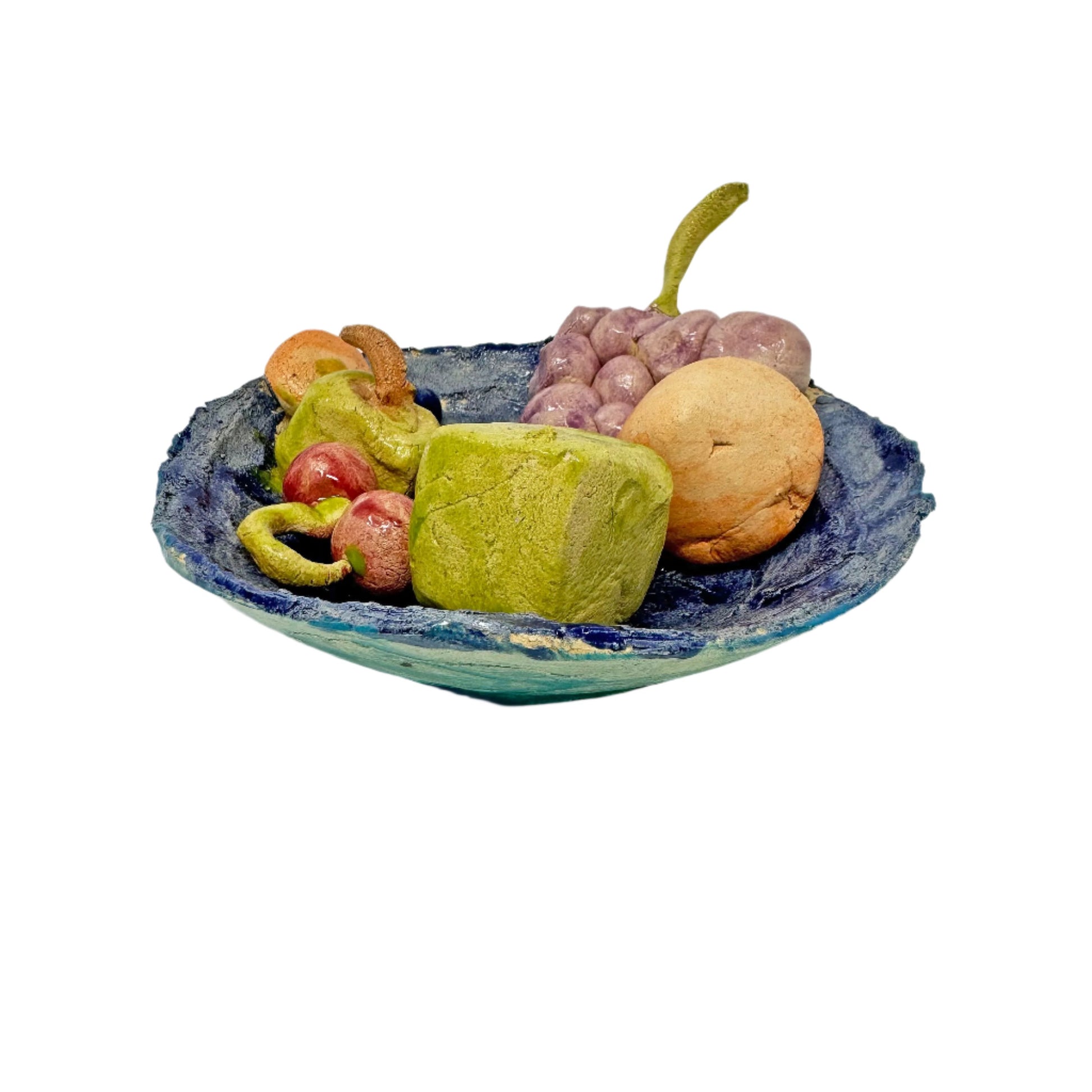 A glazed ceramic sculpture of a bowl of fruit, photographed from the side. The bowl is painted in shades of light and dark blue. The bowl contains hand-built ceramic forms representing purple grapes, red cherries, an orange, a green apple. The is also a large green cube in the bowl. 