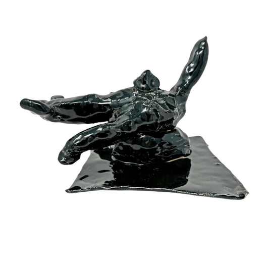 A small ceramic sculpture coated with shiny black glaze. The object is abstract and represents a cat positioned on a flat square base. The body of the animal is two stacked rounded shapes, with long limbs including a tail extending outward. 