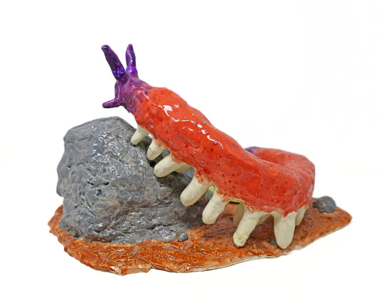 A ceramic caterpillar-like figure with a pink body, white feet, and purple head. The figure is crawling up a gray rock, which sits atop a brown base. 