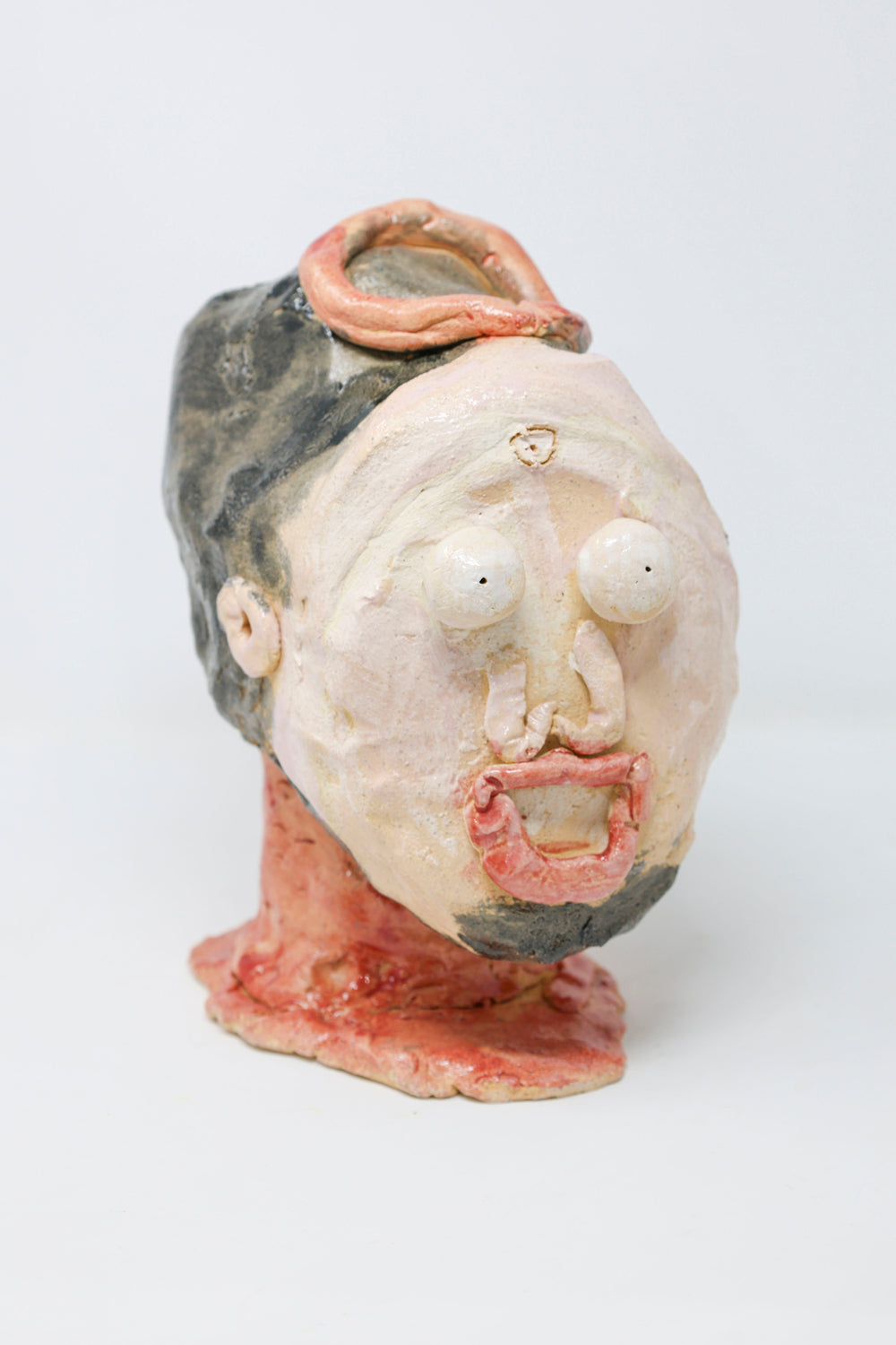 Ceramic sculpture of a head with features expressing fright. The figure has black hair and a goatee, a red neck and is wearing circular red crown. 