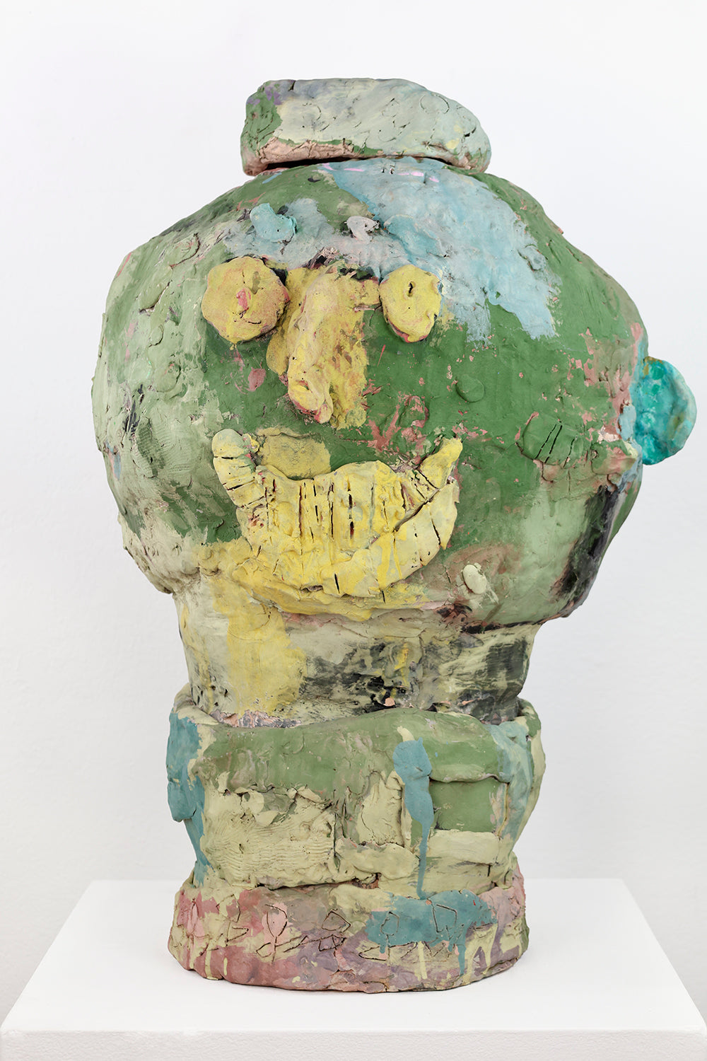 Green ceramic sculpture of a head against a white background. The facial features are yellow. The face is smiling, and has patches of blue and lighter shades of green scattered throughout. 