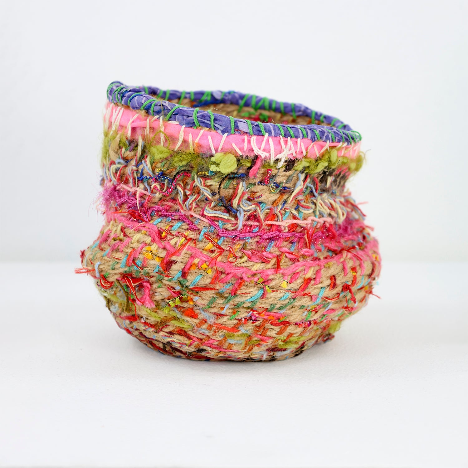 A colorful, handcrafted basket with a cylindrical shape and a textured surface. It is intricately woven with a variety of materials, including yarn and fibers in vivid hues.  The body of the basket is woven with natural and bright colors, including red, pink, blue, and yellow, creating a patchwork effect. 