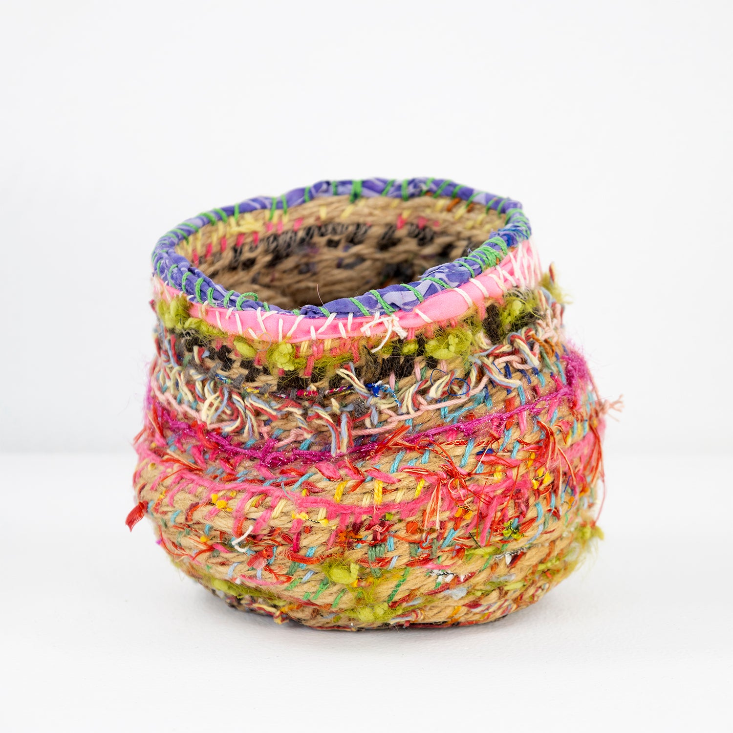 A colorful, handcrafted basket with a cylindrical shape and a textured surface. It is intricately woven with a variety of materials, including yarn and fibers in vivid hues.  The body of the basket is woven with natural and bright colors, including red, pink, blue, and yellow, creating a patchwork effect. 