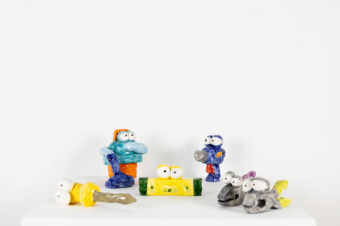 A selection of colorful ceramic sculptures arranged on a white pedestal. The objects are playful characters shaped like tools you might find in a wood shop: a screwdriver, pliers, a vacuum, a drill, and a level. Each item is personified with two large round eyes and a distinct expression.