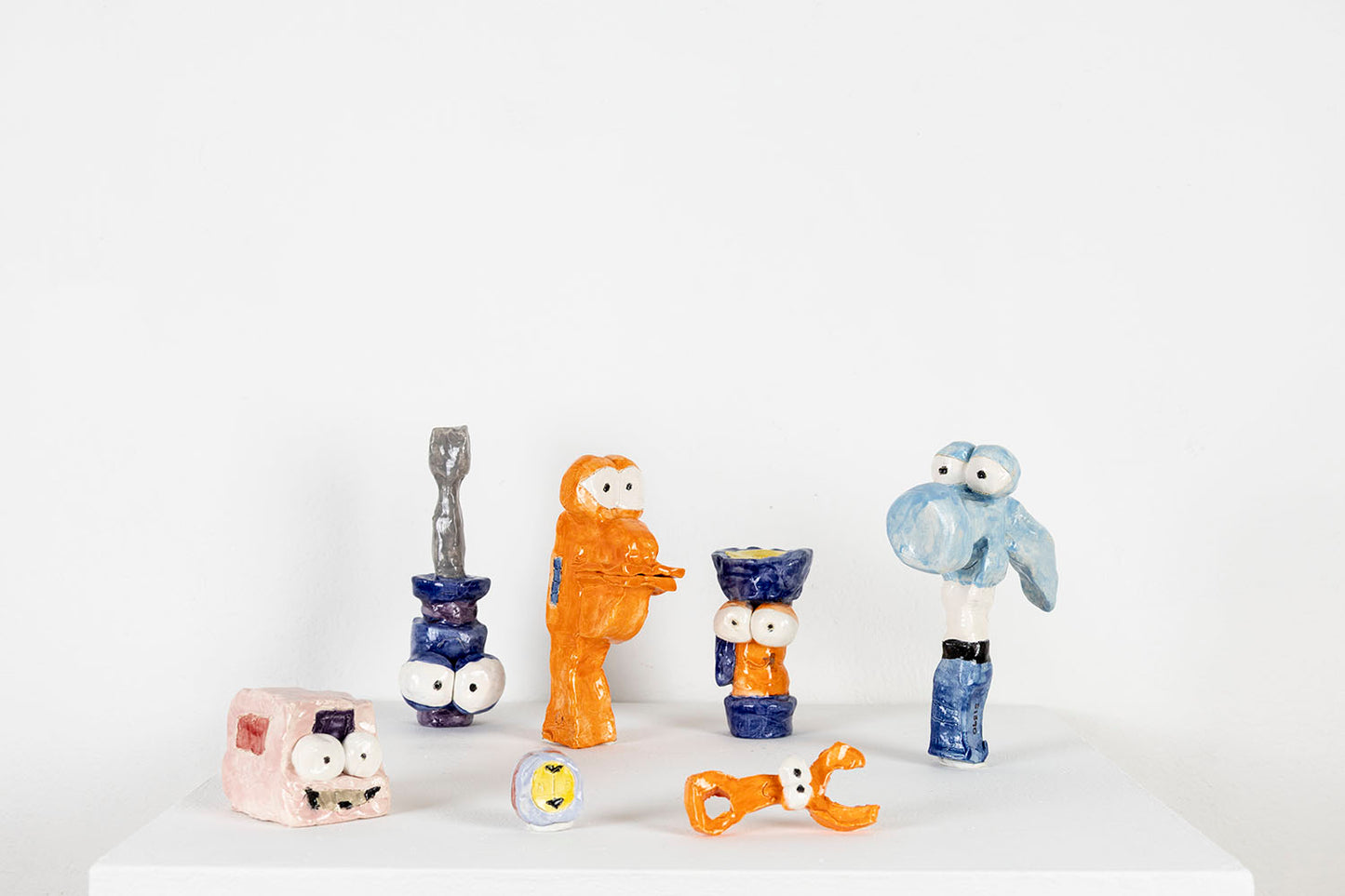 A selection of colorful ceramic sculptures arranged on a white pedestal. The objects are playful characters shaped like tools you might find in a wood shop: a screwdriver, a hammer, a wrench, and more. Each item is personified with two large round eyes and a distinct expression.