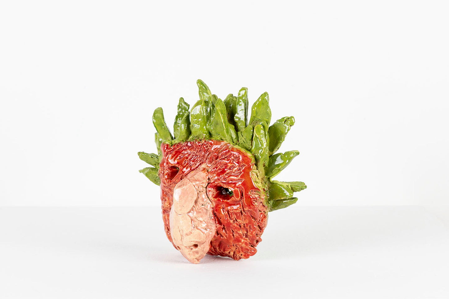 Photographed facing the left: a small ceramic mask of a human-like face with a large snout. The mask is glazed in glossy red, pink, and green. The surface of the face is red and textured with ragged incised striations. A green crown of smooth pointed leaves fans out around the top of the head. 