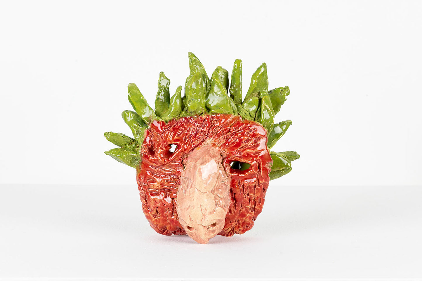 A small ceramic mask of a human-like face with a large snout, photographed from the front. The mask is glazed in glossy red, pink, and green. The surface of the face is red and textured with ragged incised striations. A green crown of smooth pointed leaves fans out around the top of the head. From this angle, the background is visible through the eye holes. 