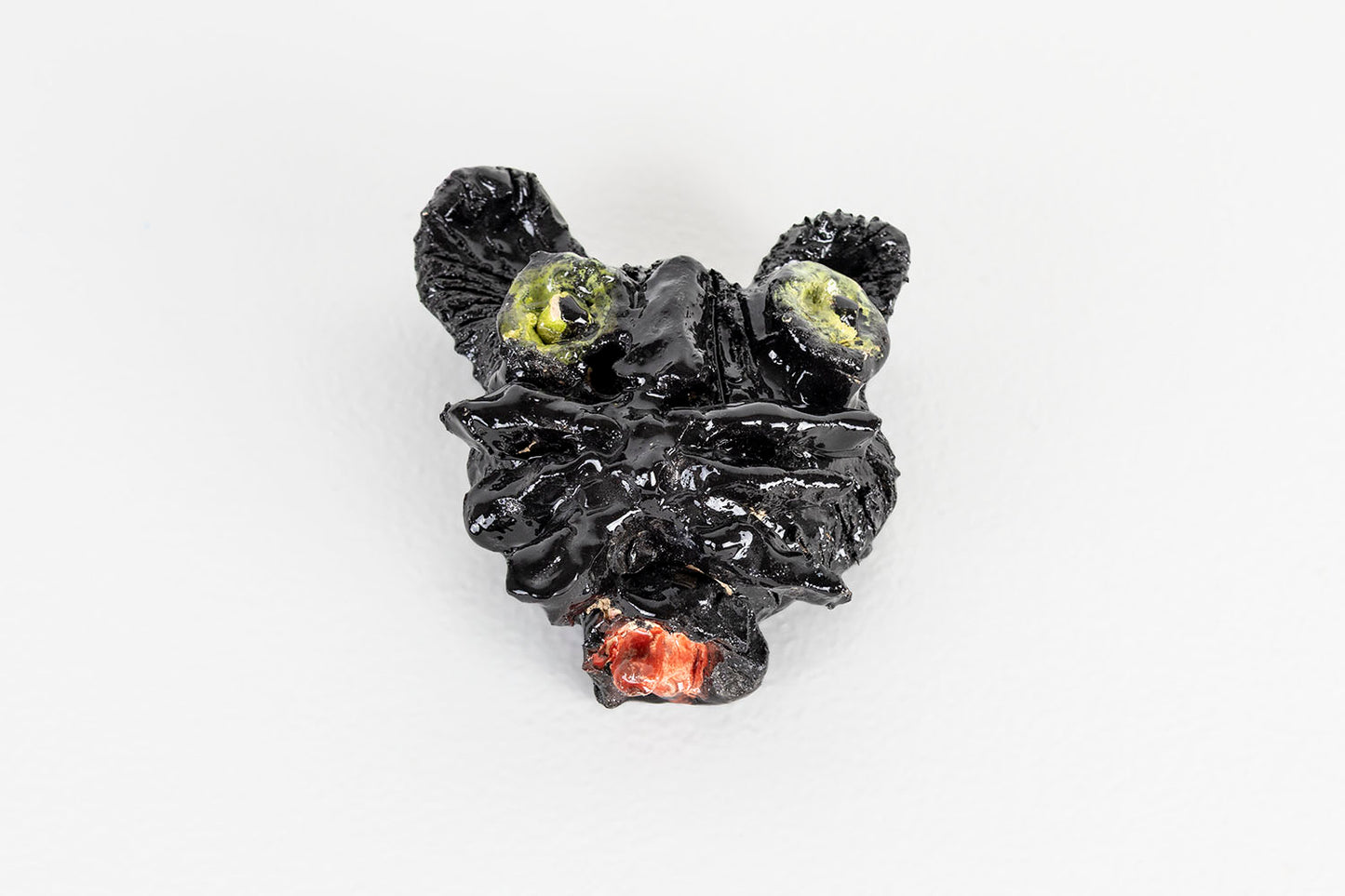 A ceramic mask shaped like a black cat's head. The cat has big round green eyes, long black whiskers, and a bright red tongue protruding from a round mouth. 