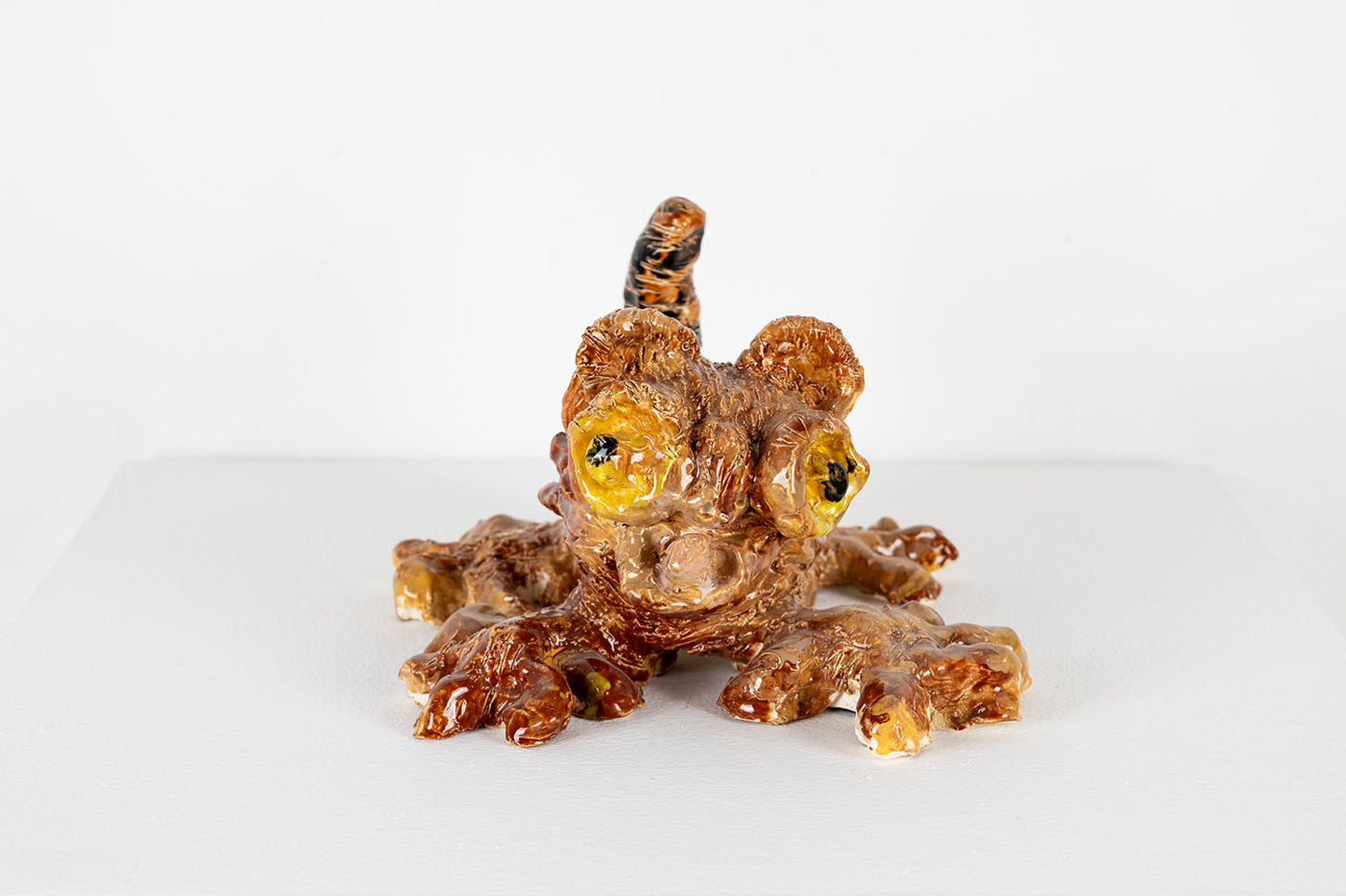 A small ceramic sculpture of a brown and black kitten. All four of its limbs are splayed out in a playful position, and its pointed tail sticks straight up. It has wide yellow eyes and a little pointy tongue protruding from its round mouth. 