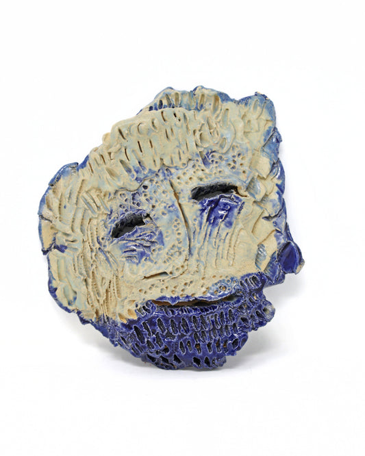 This is ceramic sculpture is an abstract mask. The facial features are carved out and highlighted with a dark blue glaze. The rest of the face being a beige and light blue glaze. 