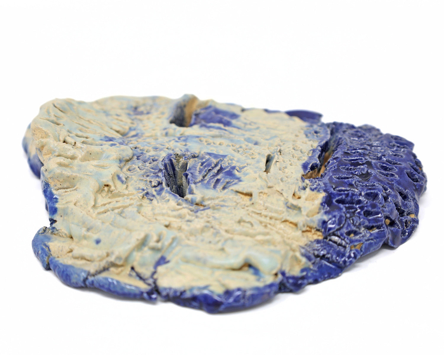 This is ceramic sculpture is an abstract mask. The facial features are carved out and highlighted with a dark blue glaze. The rest of the face being a beige and light blue glaze. 