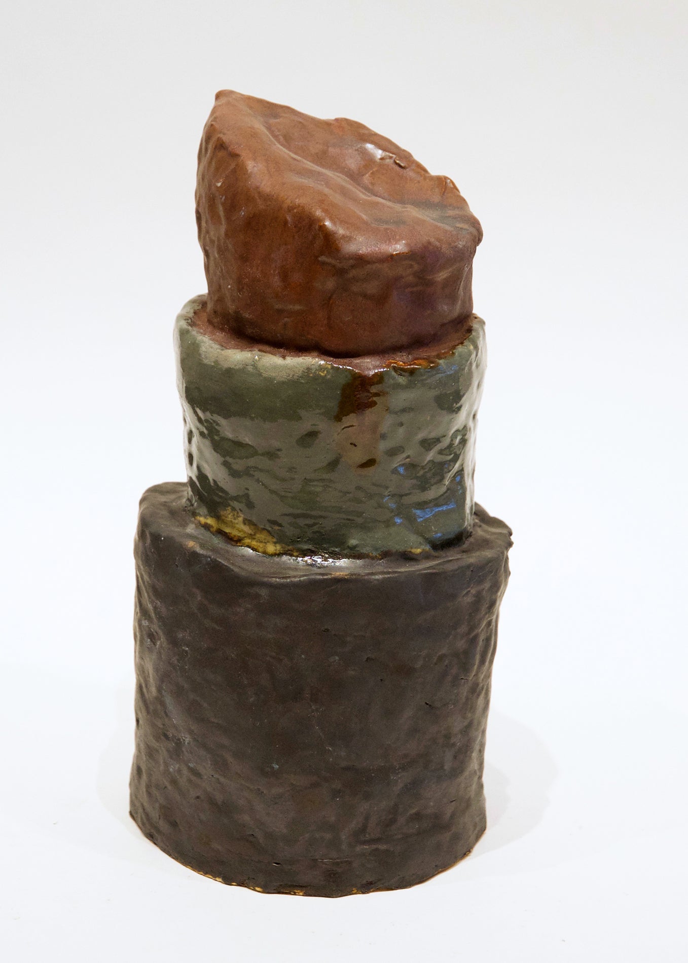 A ceramic sculpture representing a larger-than-life tube of lipstick. The sculpture is three stacked cylinders, the bottom one glazed a deep brown, the center a shiny olive green, and the top a warm reddish brown. 