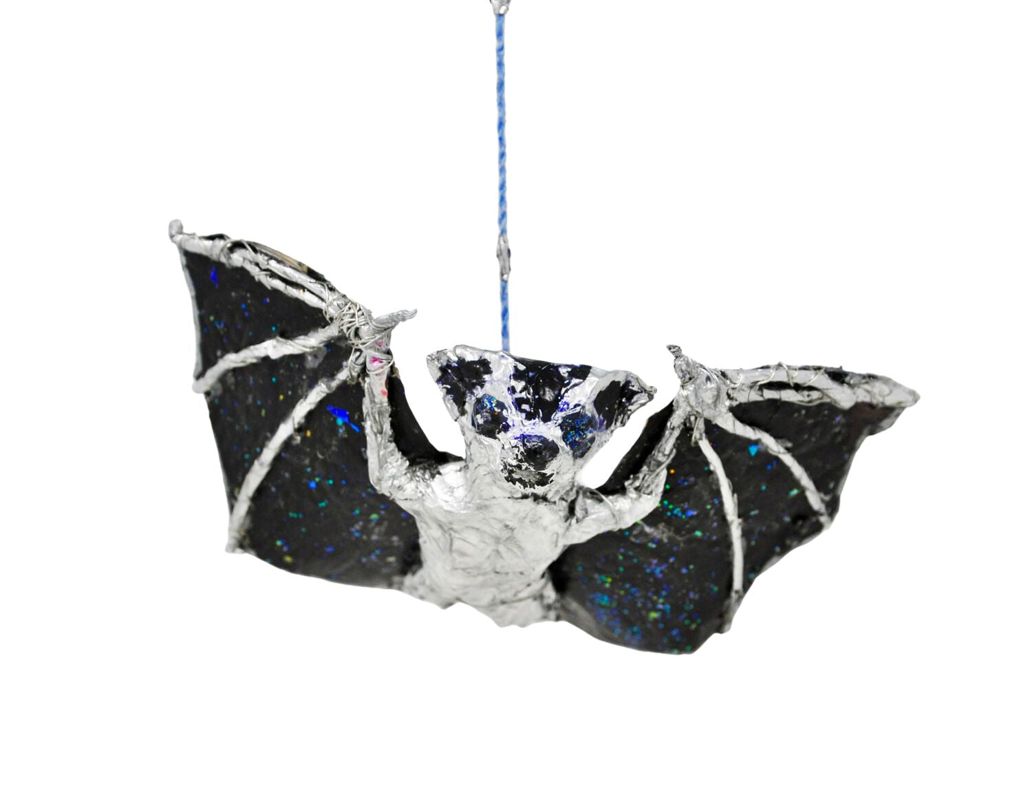 a hanging ornament shaped like an animal: a bat in flight. The bat is made with various media including wire, tape, glitter, paint, and other recyclable materials. The body of the bat is painted silver, and the wings are painted black with blue and green glitter sprinkled over the surface. The object hangs from a twisted blue and periwinkle thread. Photographed from this angle, the bat is flying straight toward the camera, with black eyes and little pointed ears. 