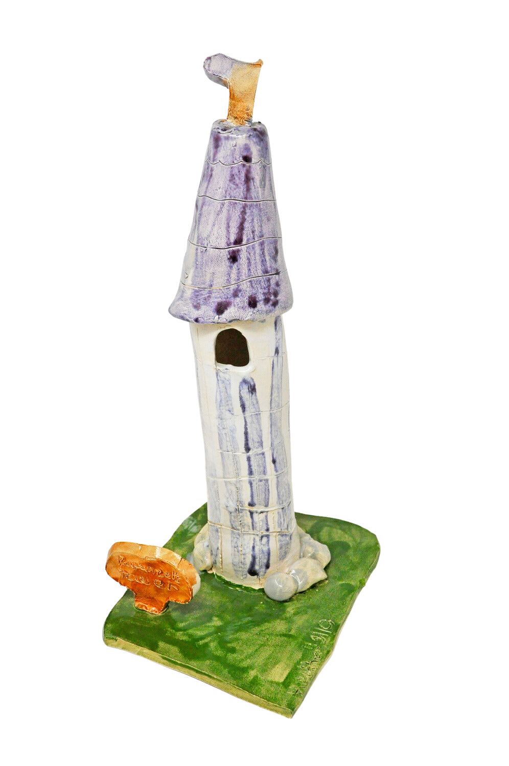 a ceramic sculpture of a white and purple medieval tower with one window and a little flag on the top. It is sitting on a green lawn next to a little brown ceramic sign with the words "Rapunzel's Tower."