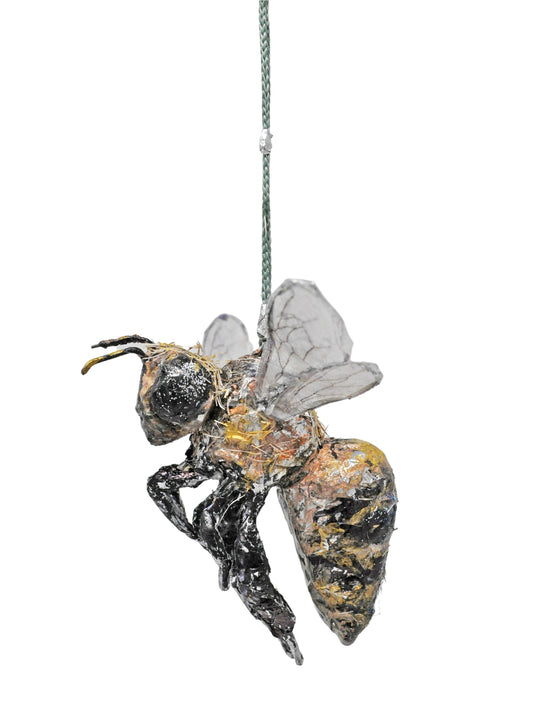 a hanging 3-dimensional ornament in the shape of a realistic bumble bee. The object was made using various media, including wire, tape, paint, thread, and recyclable plastics and paper material. The bee is painted to have a black and yellow striped thorax, black legs, large black eyes, and wire wings filled with clear glue or plastic. The object hangs from a twisted green and yellow thread. In this photograph, the bee is in profile, facing toward the left. 