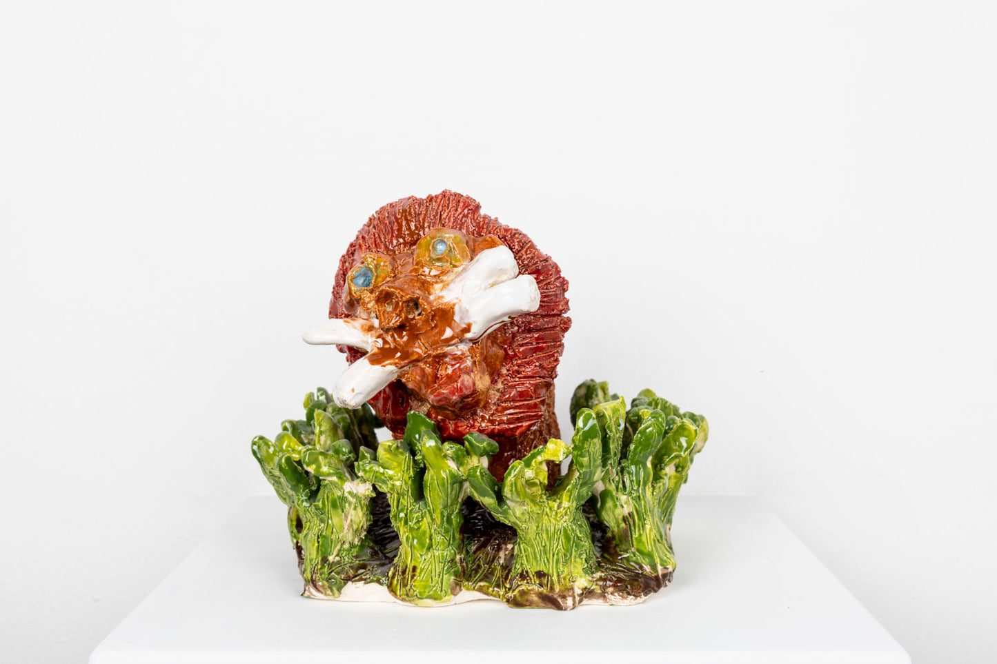 Glazed ceramic sculpture of a reddish orange lion's head with bared white whiskers emerging from a bed of green ceramic grass. 