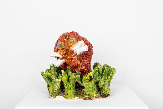 Glazed ceramic sculpture of a reddish orange lion's head with bared white whiskers emerging from a bed of green ceramic grass. 