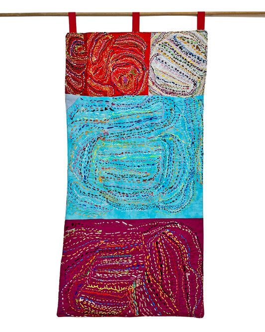 An intricately embroidered textile wall-hanging, hanging from a wooden dowel from three red loops. The work is a long rectangle made up of 5 squares of fabric. The central section is a light blue color, with smaller red, grey, and maroon squares stitched above and below. Over the entire surface of each fabric swatch, swirling lines and stripe designs are embroidered in running stitches using every color of the rainbow. 