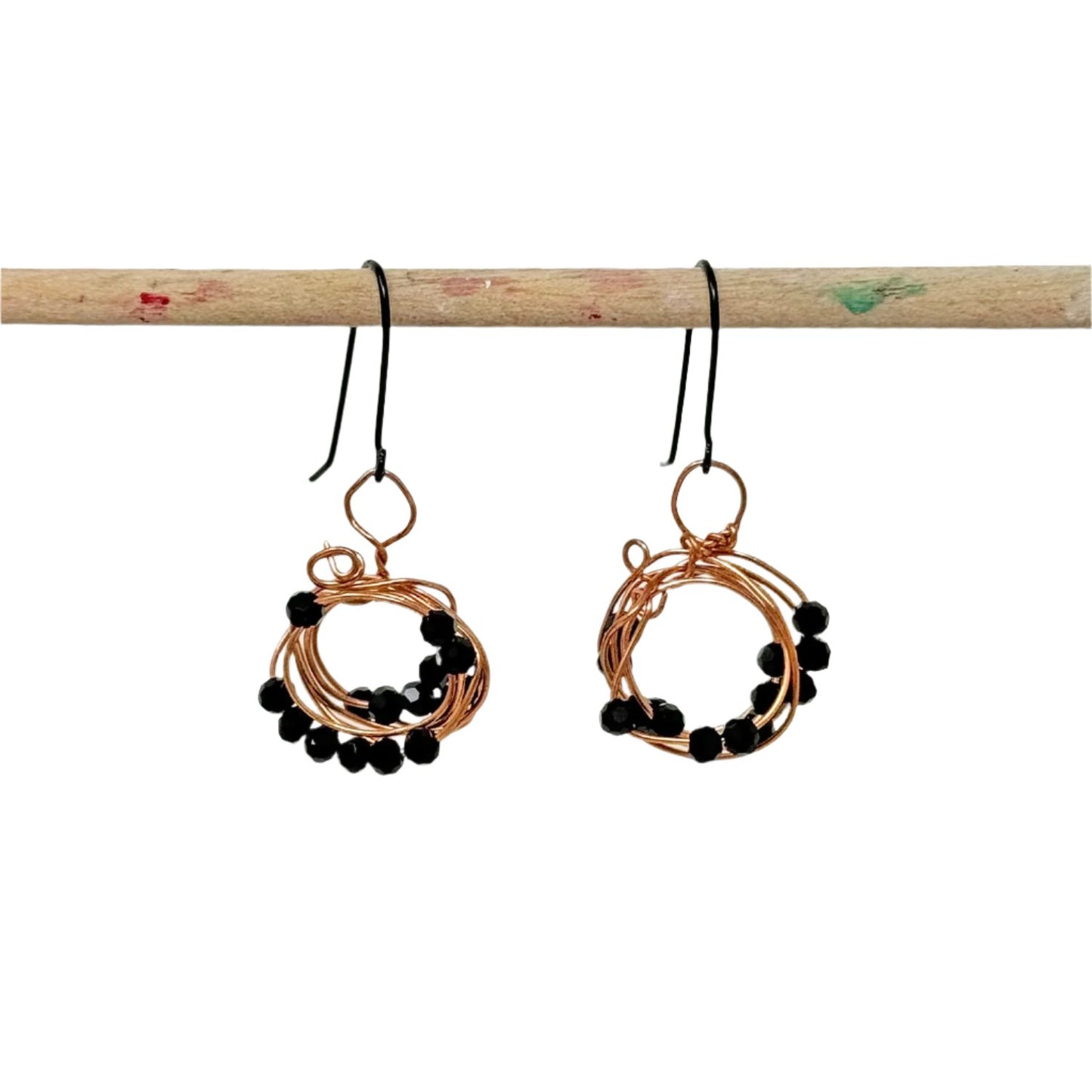 A set of unique handmade earrings hanging from a wooden dowel. The earrings are made from coiled copper wire and black beads. The design of each earring is concentric wire circles that have been sparsely strung with octagonal crystal beads. 