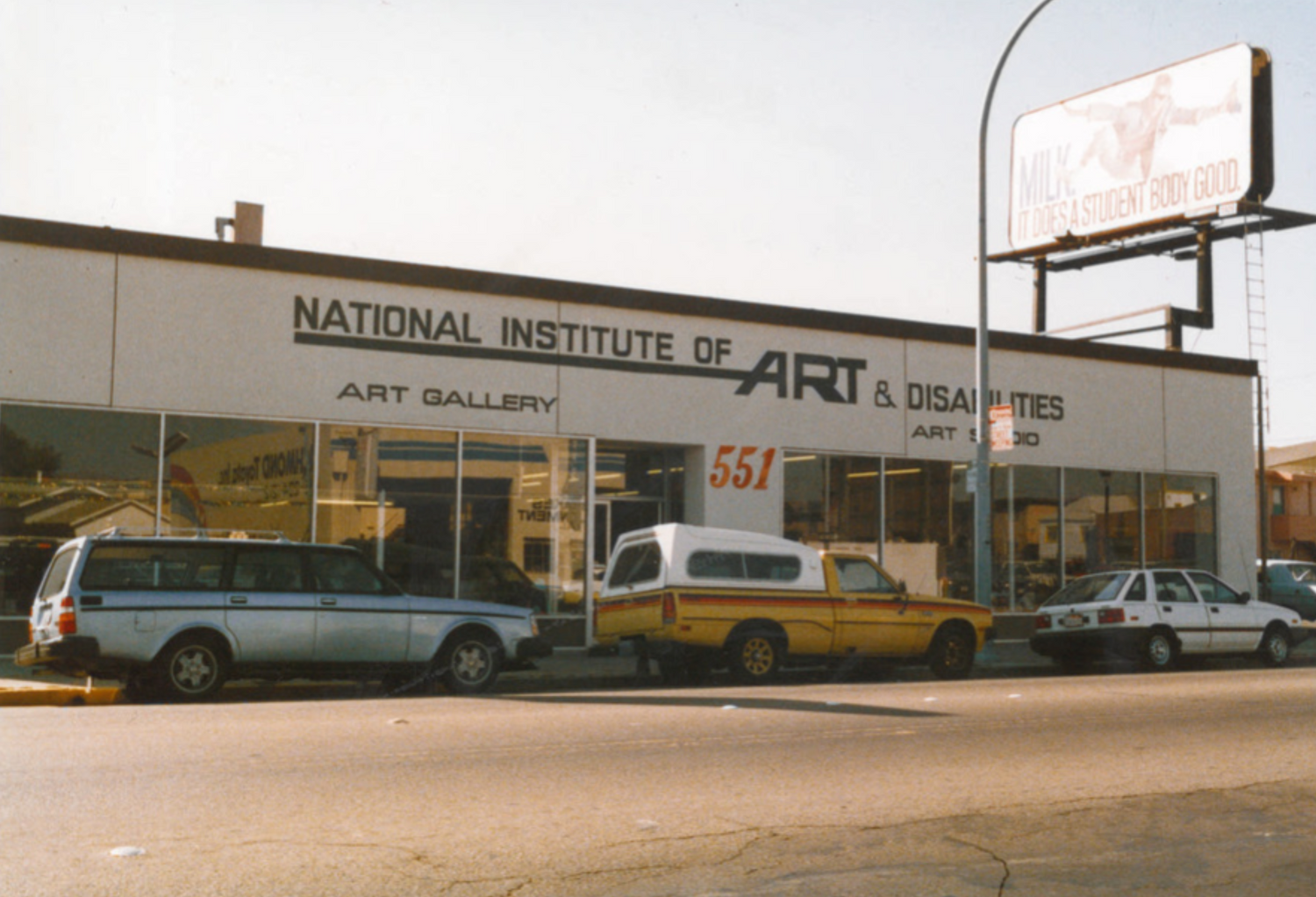 An image from the early 1980s of the front of a building, photographed from across the street on a sunny day. The facade of the building is white with hand-painted black text that reads "National Institute of Art & Disabilities: Art Gallery, Art Studio." The building is a storefront with a row of tall dark windows. There are 3 cars parked in front of the building, and a billboard advertising milk on the roof. 