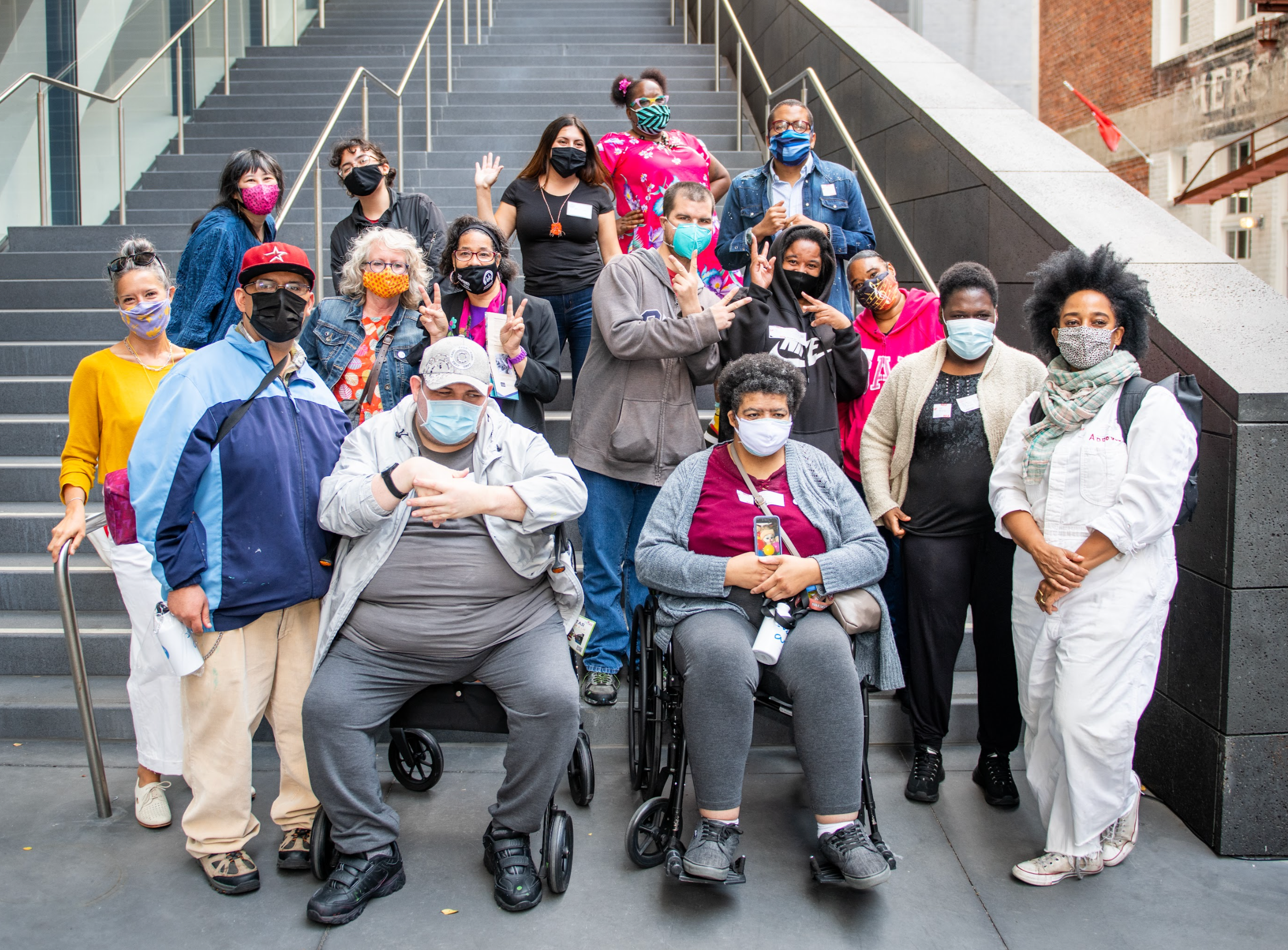 A photograph of a group of people gathered in front of a tall set of grey stone stairs.  The people are all wearing face masks and looking at the camera. Some of the people are seated in wheelchairs. Some are waving at the camera or holding up peace signs. 