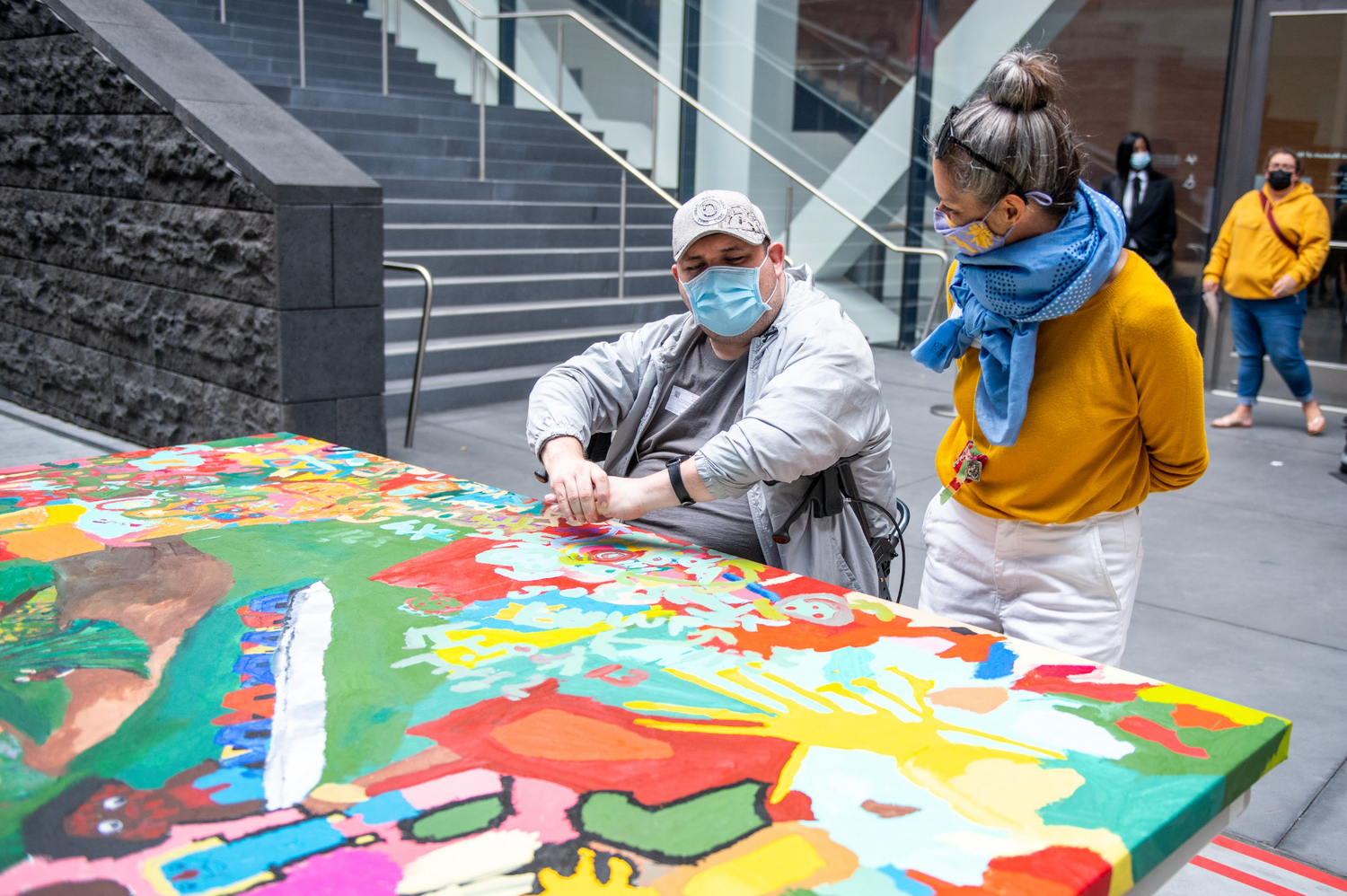 A photograph of two people in a museum courtyard working on a large painting. The figure in the center is man with light skin who is seated in a walker. He wears a grey baseball cap, a blue face mask, and a grey jacket. A white woman with grey hair in a bun stands to him. She is wearing a blue scarf, yellow sweater, and white pants.  A large painting with green, yellow, and red shapes is laid out flat on a table in front of them. 