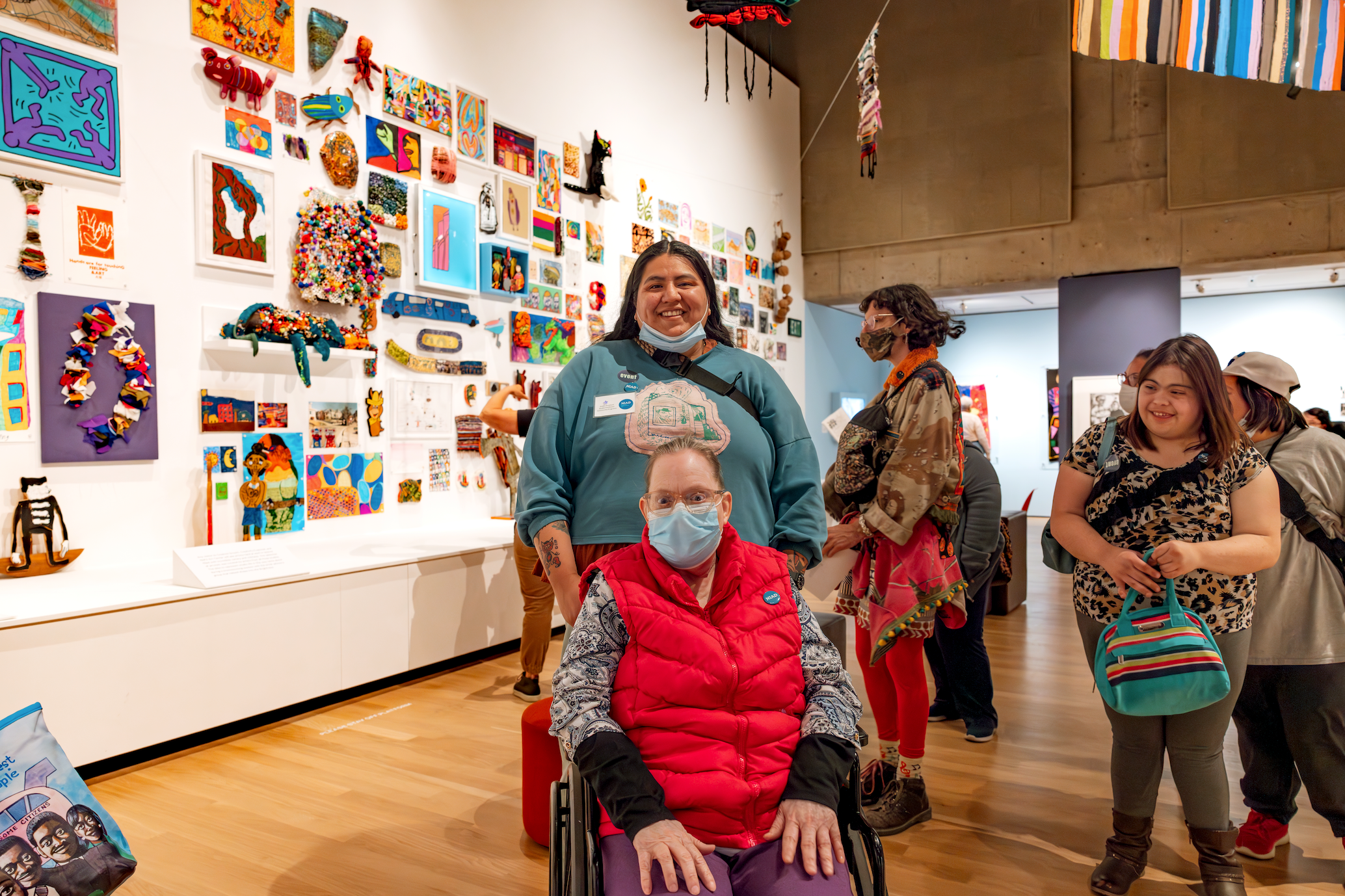 A group of people in a museum gallery. In the foreground there is a figure seated in a wheelchair, looking at the camera and wearing a bright red vest, purple pants, and a blue face mask. Behind them, a figure stands smiling at the camera, with long black hair and a blue sweatshirt. The wall to the left is hung salon style with colorful artworks made in every medium, arranged in a large grid. A group of people stand in the space, admiring the art. 