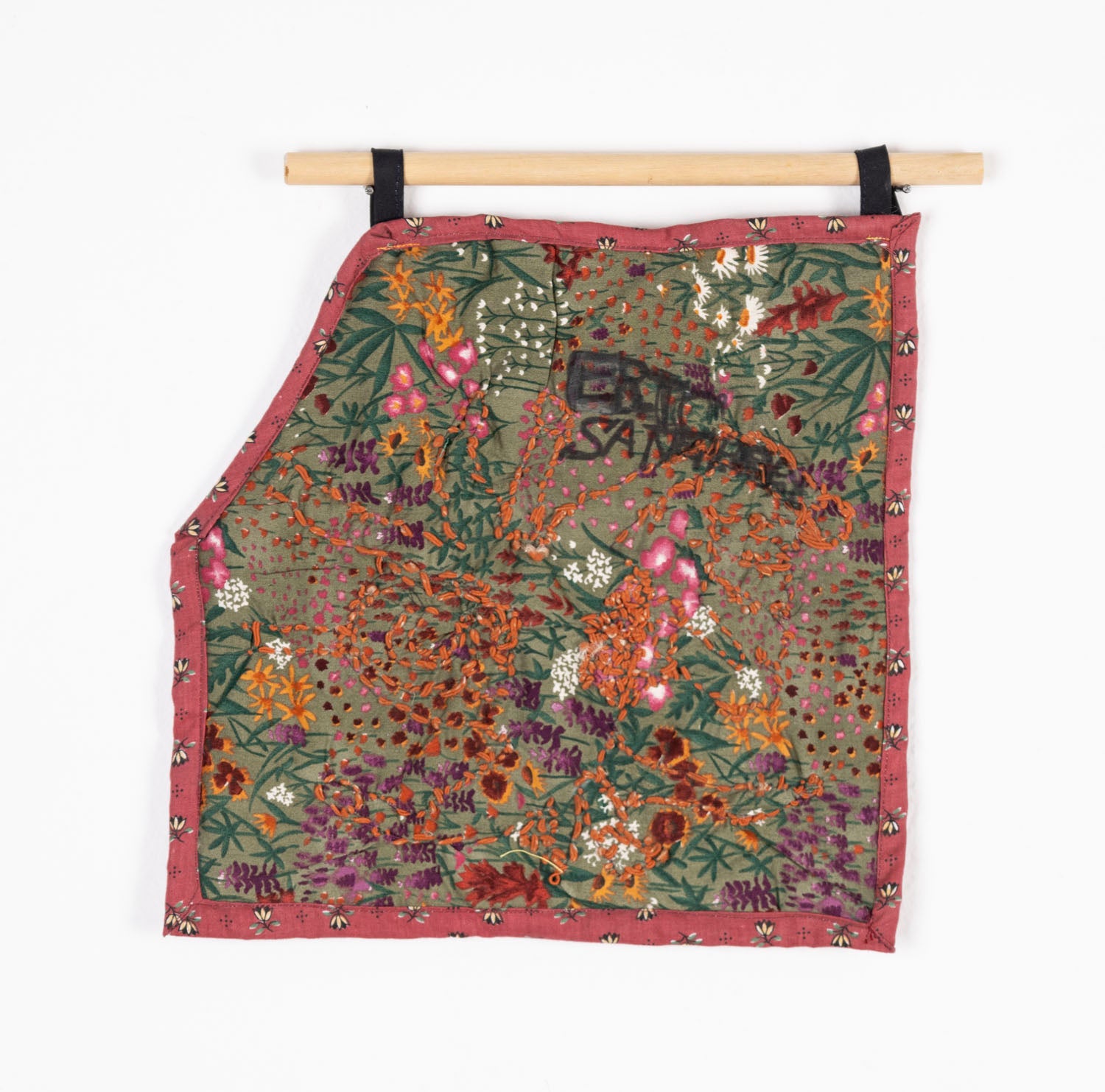 A shaped wallhanging textile work hanging from a wooden dowel and photographed against a white backdrop. The main swatch of fabric is printed with an image of a garden: pale green ground with yellow, white, red, and purple flowers. The fabric is cut like a square with one corner folded down. Over this design, the artist has embroidered a swirling shape using orange thread and written words including "Eric" with black fabric marker. The piece has a dark pink floral fabric border.