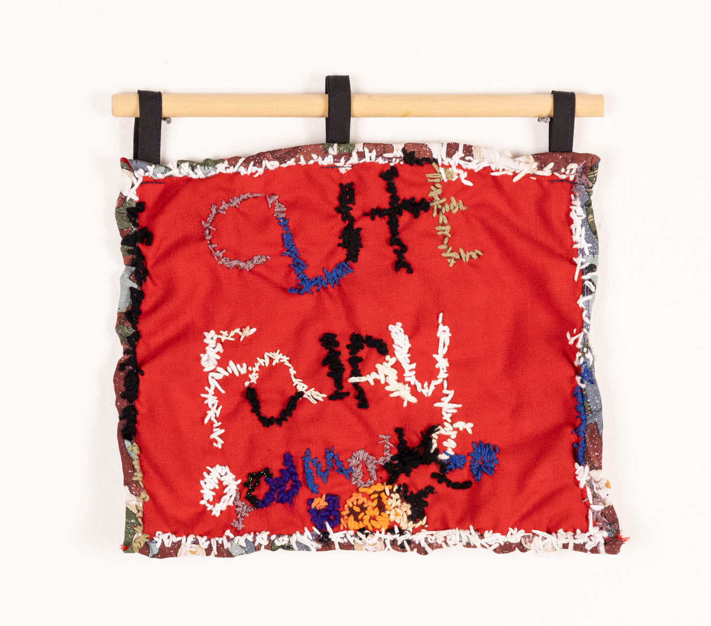 A square textile piece hanging from a wooden dowel. On a bright red swatch of fabric, the artist has embroidered the words "cute fairy godmother" using red, white, and black embroidery thread. The red fabric is stitched to a brown, green, and grey piece of fabric which creates the backing and thin border. 