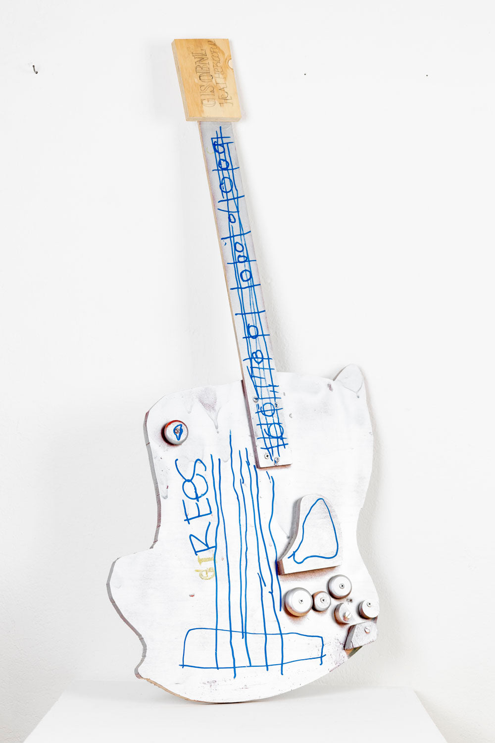 A mixed media sculpture in the shape of an instrument. Several thin pieces of wood have been cut to mimic the body and bridge of an electric guitar, with metal screws and beads adhered as tuning knobs. The sculpture is painted all white and the frets and strings are drawn with bright blue paint marker. The artist has added hand lettered text in pencil and marker. 