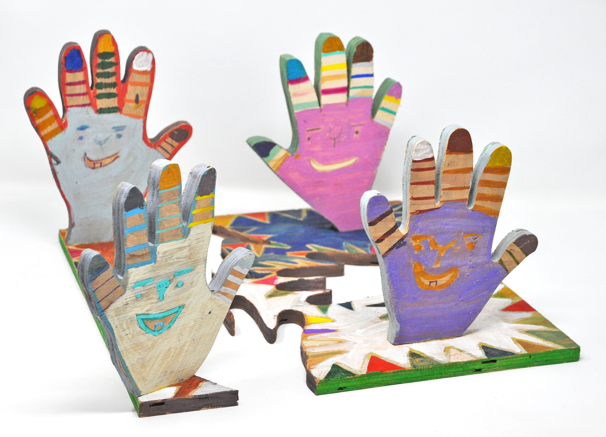 A wooden sculpture of 4 hands standing perpendicular to a wooden base from which they were cut out of. Each hand is a different color, white, light blue, lilac, and pink, and each has a smiling face on the palm. The fingers are painted in different colored stripes. 