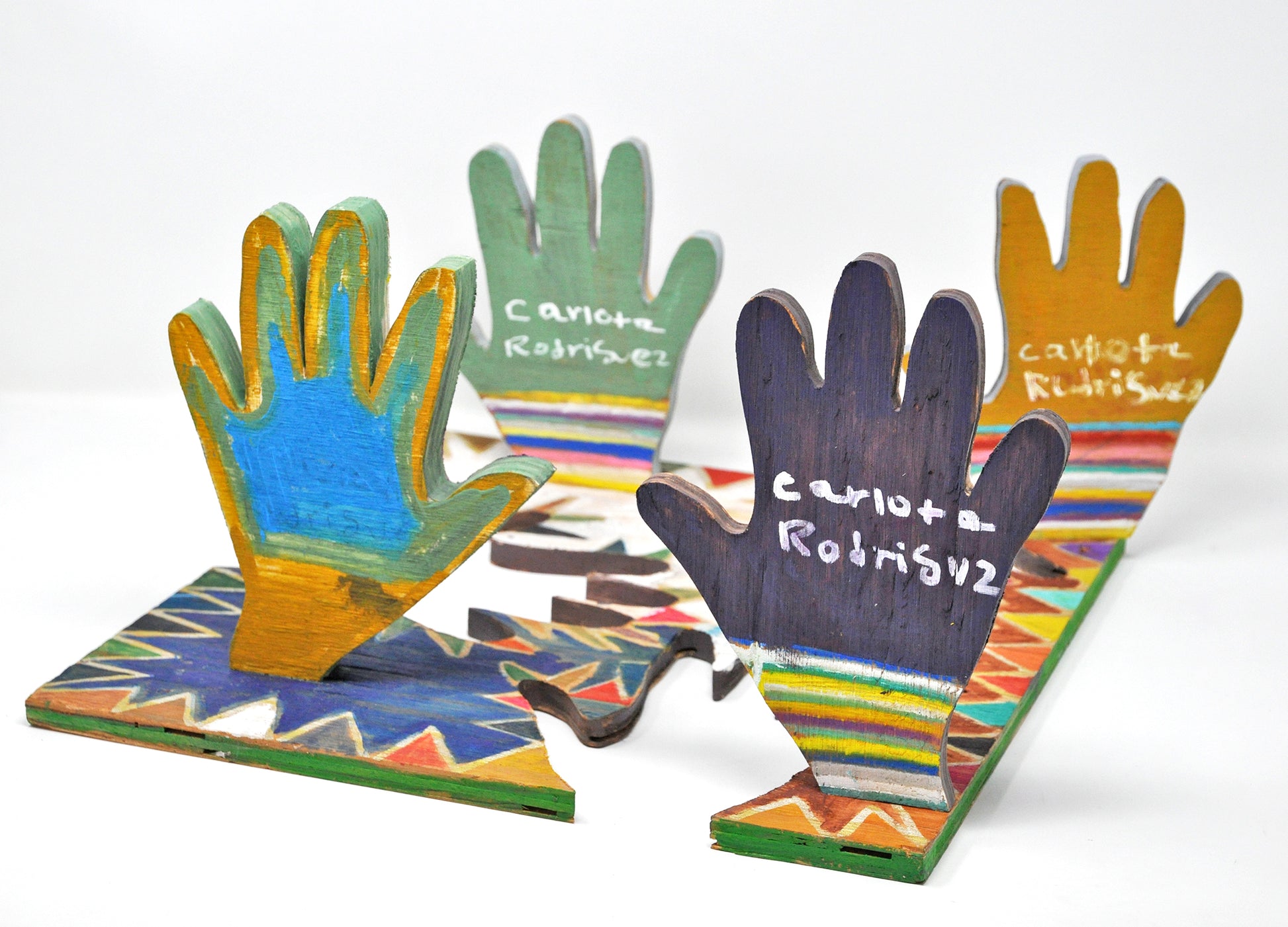 Photograph of the back of a wooden sculpture with 4 colorful hands standing perpendicular to a wooden base from which they were cut out of. Three of the four hands have the artist's signature written in white on them, and one has her name written in pencil, obscured by paint on top of it. 