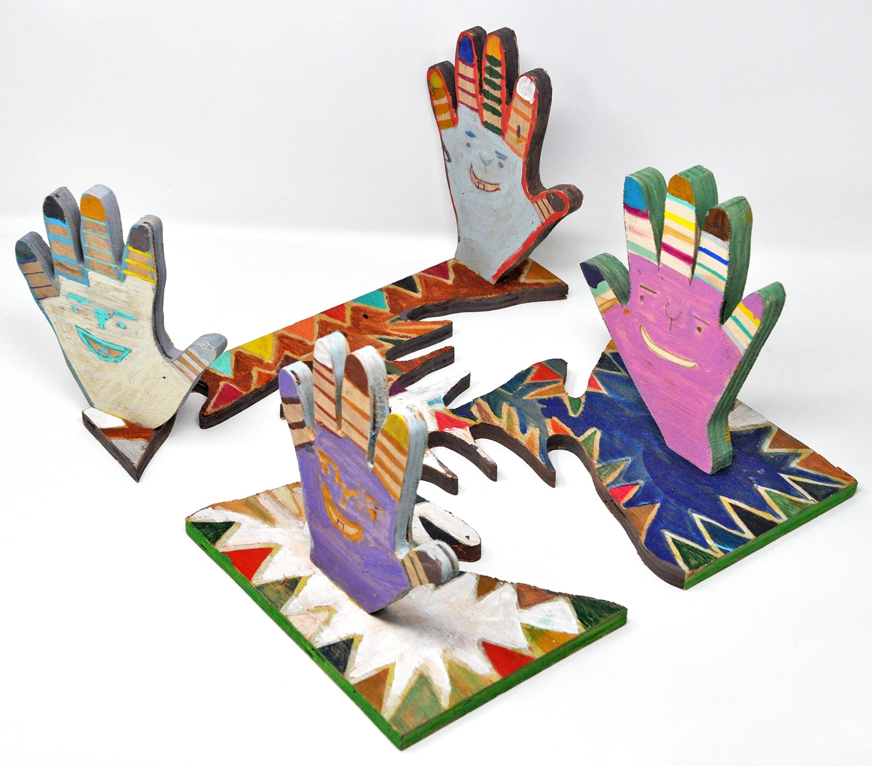 A wooden sculpture of 4 hands standing perpendicular to a wooden base from which they were cut out of. Each hand is a different color, white, light blue, lilac, and pink, and each has a smiling face on the palm. The fingers are painted in different colored stripes. 