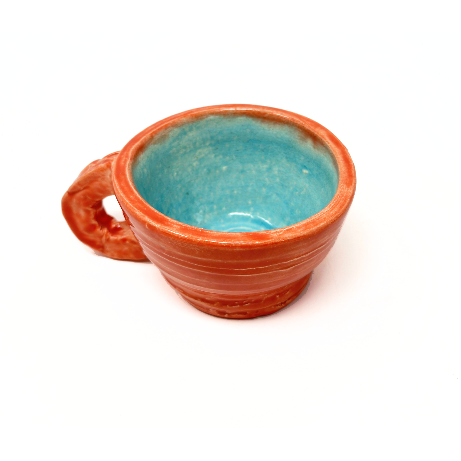 A ceramic mug with a small round handle and a wide circular rim. The exterior of the mug is painted a warm coral reddish orange color. The interior is glazed a light sky blue. The cup was thrown on the pottery wheel, and some of the ridges are still visible around the exterior sides. 