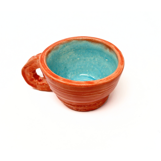 A ceramic mug with a small round handle and a wide circular rim. The exterior of the mug is painted a warm coral reddish orange color. The interior is glazed a light sky blue. The cup was thrown on the pottery wheel, and some of the ridges are still visible around the exterior sides. 